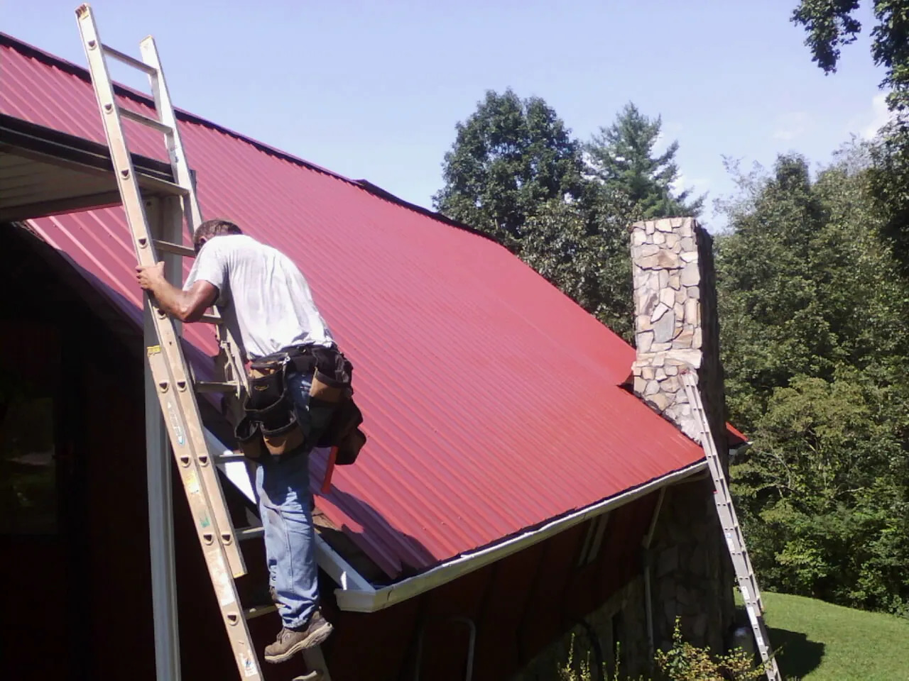 Roofer on a ladder beside a red metal roof and stone chimney — Soffit & Fascia Repair by Roofing Brothers in Norman Park